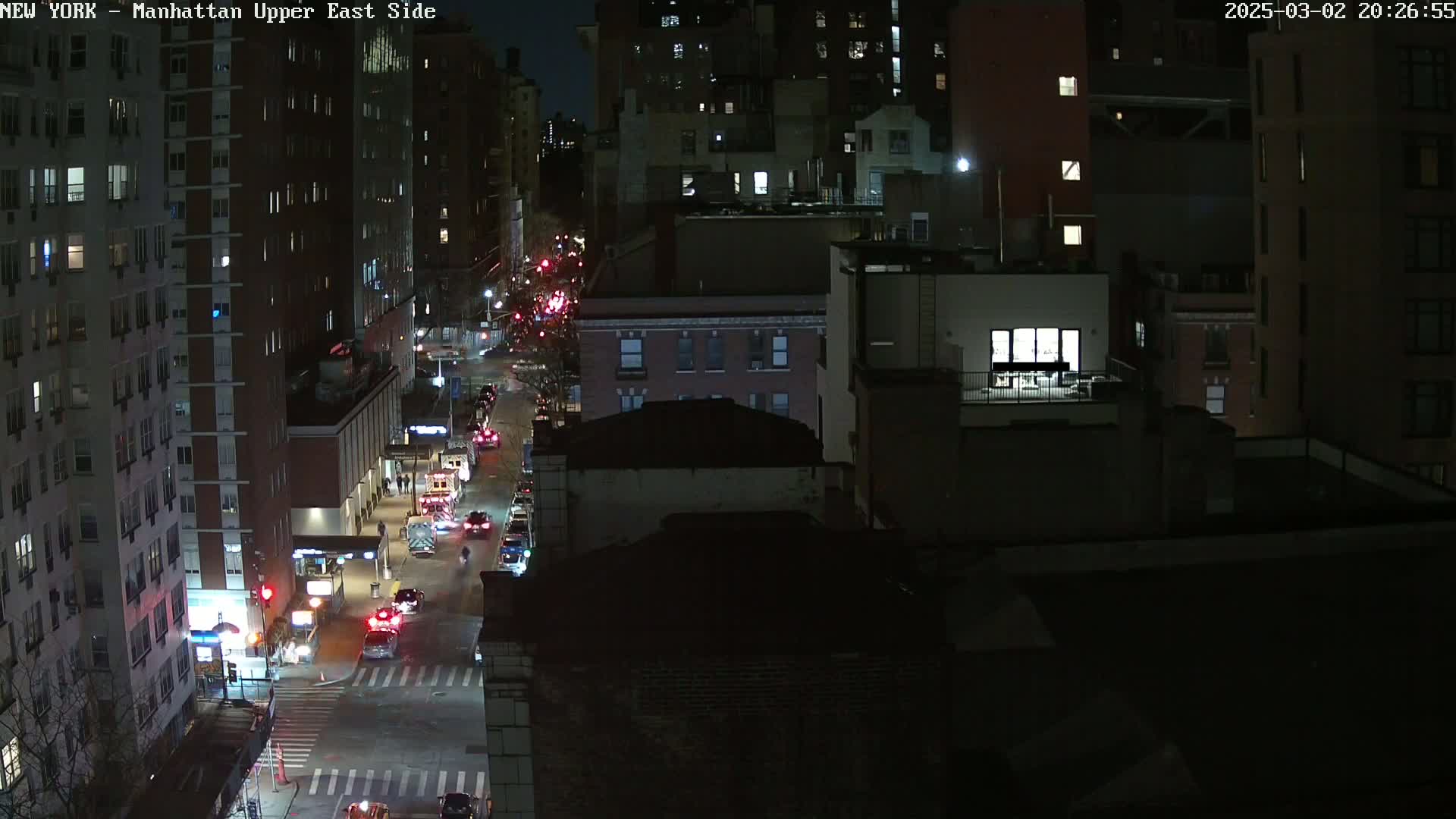 A nighttime, high-angle view of a city street with several cars driving on it, surrounded by tall buildings.
