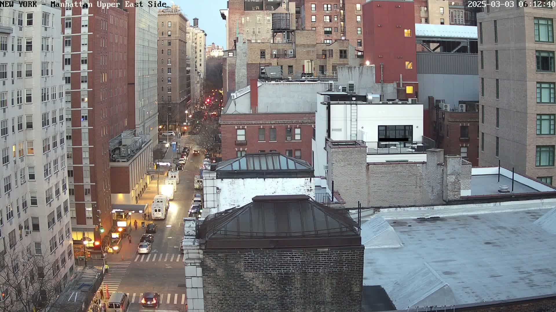 A high-angle, long shot of a city street at dusk, with several multi-story buildings lining both sides of the road and cars traveling along it.