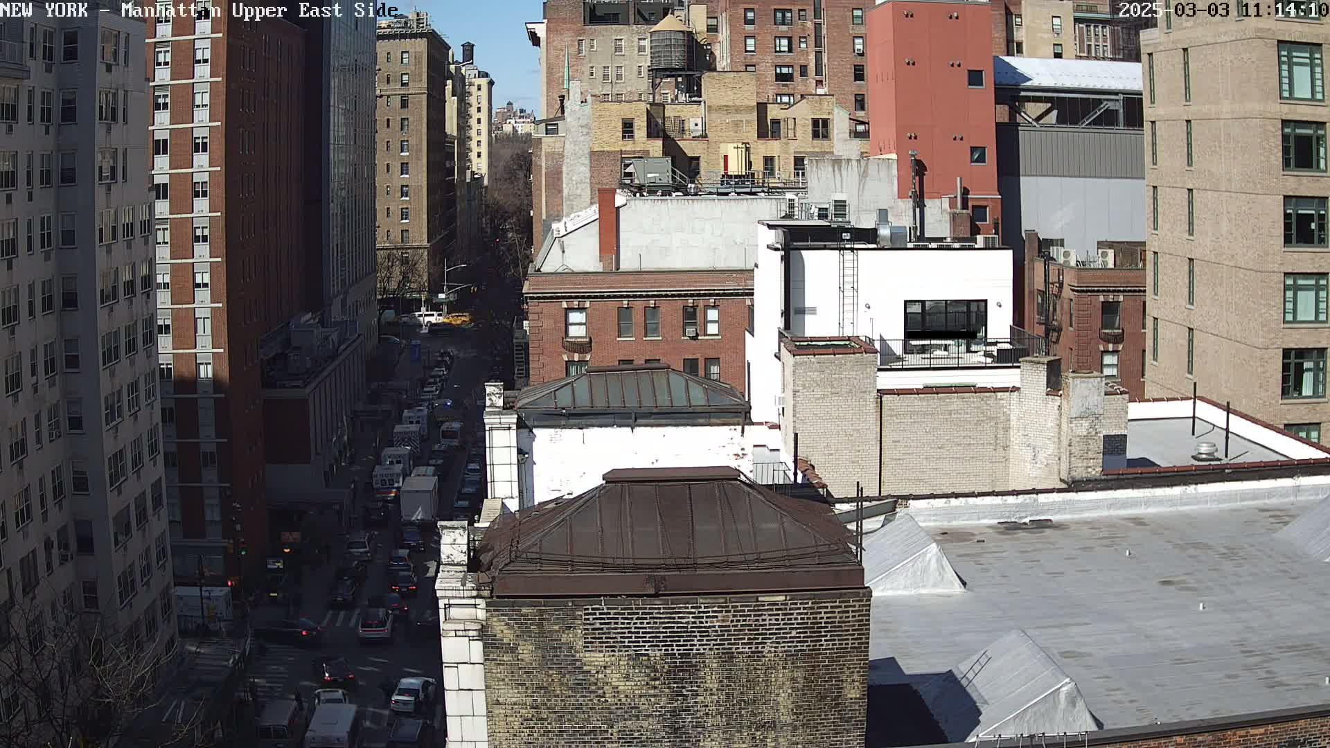A sunny day view of a city street, showing a mix of brick and stone buildings, with traffic moving along the road below.