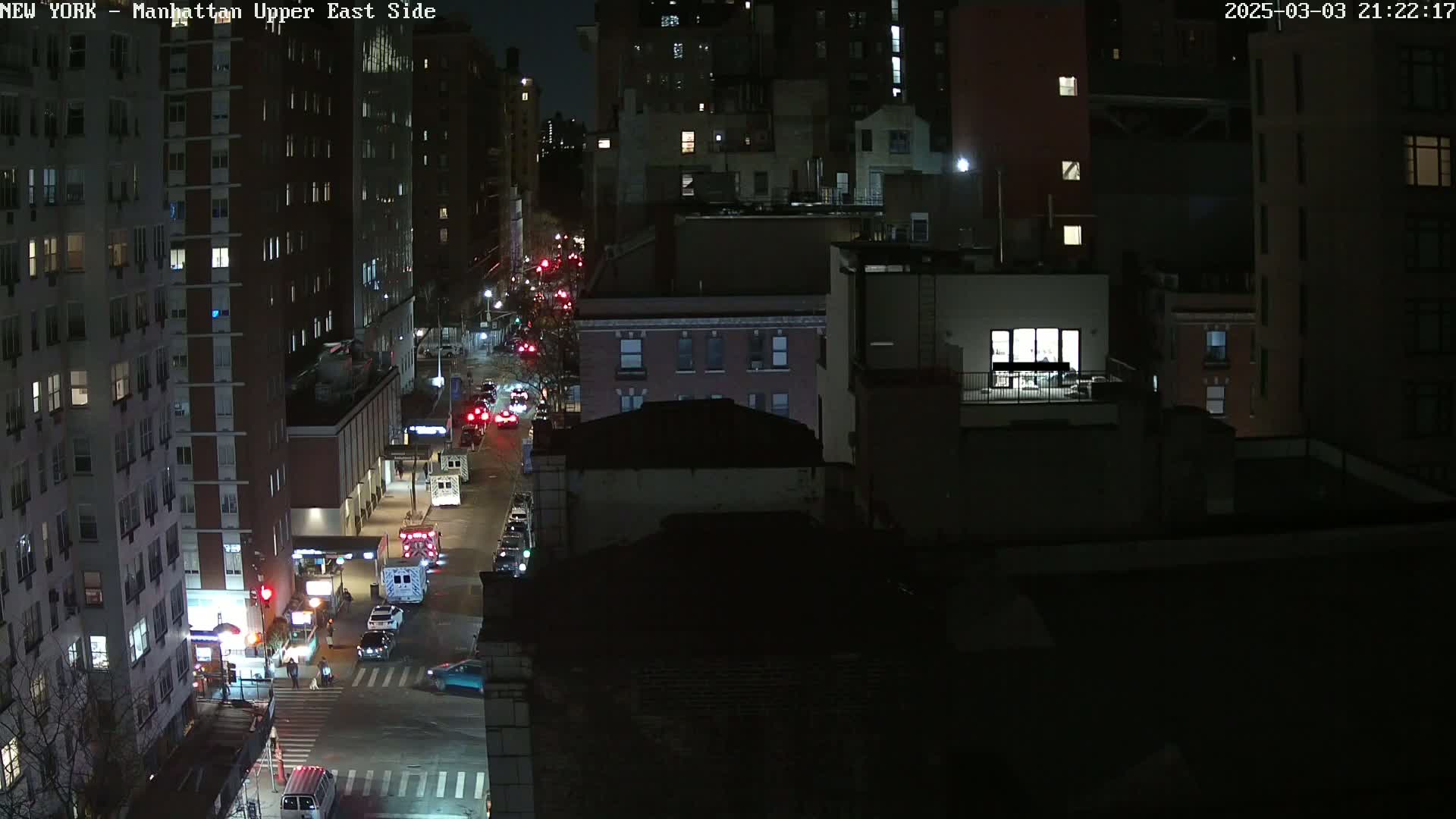 A nighttime, high-angle view of a city street with several vehicles, including emergency vehicles, and tall buildings illuminated by streetlights.
