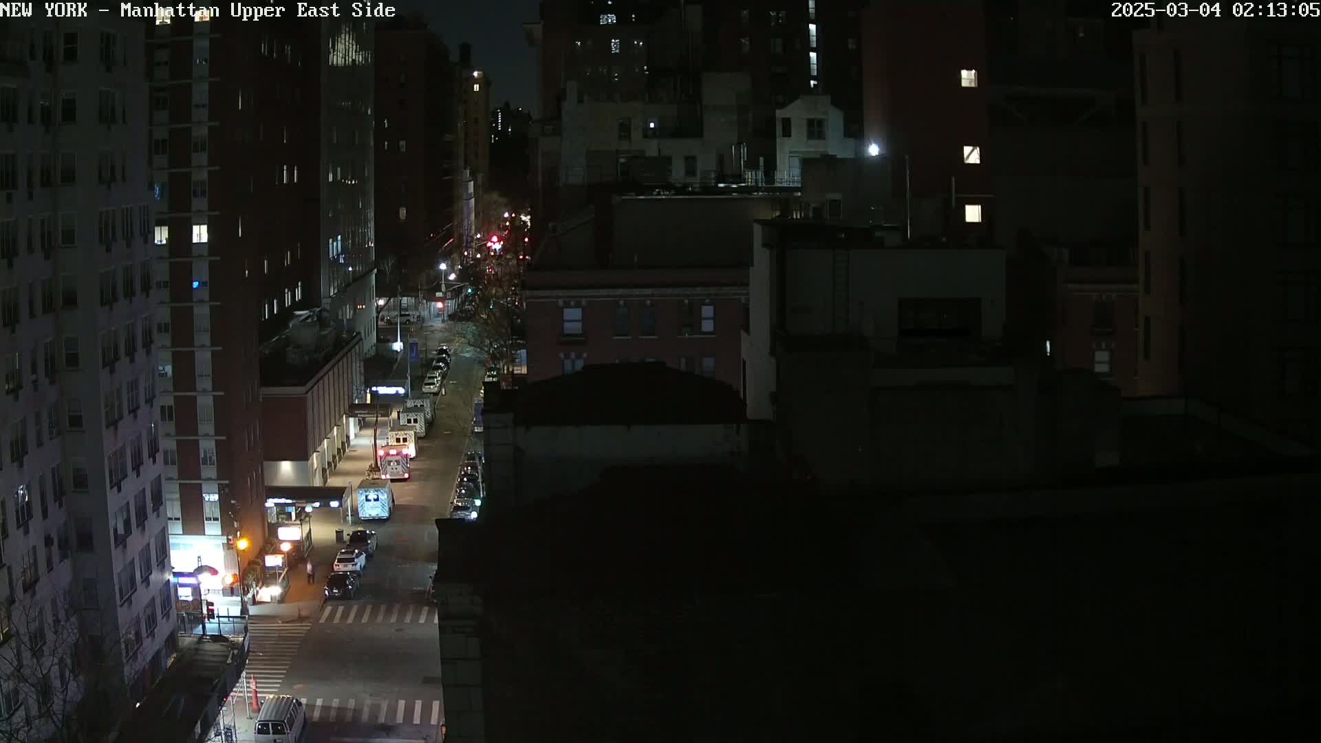 A nighttime, high-angle view shows a city street with several cars and emergency vehicles parked along the side of the road, in front of tall buildings.