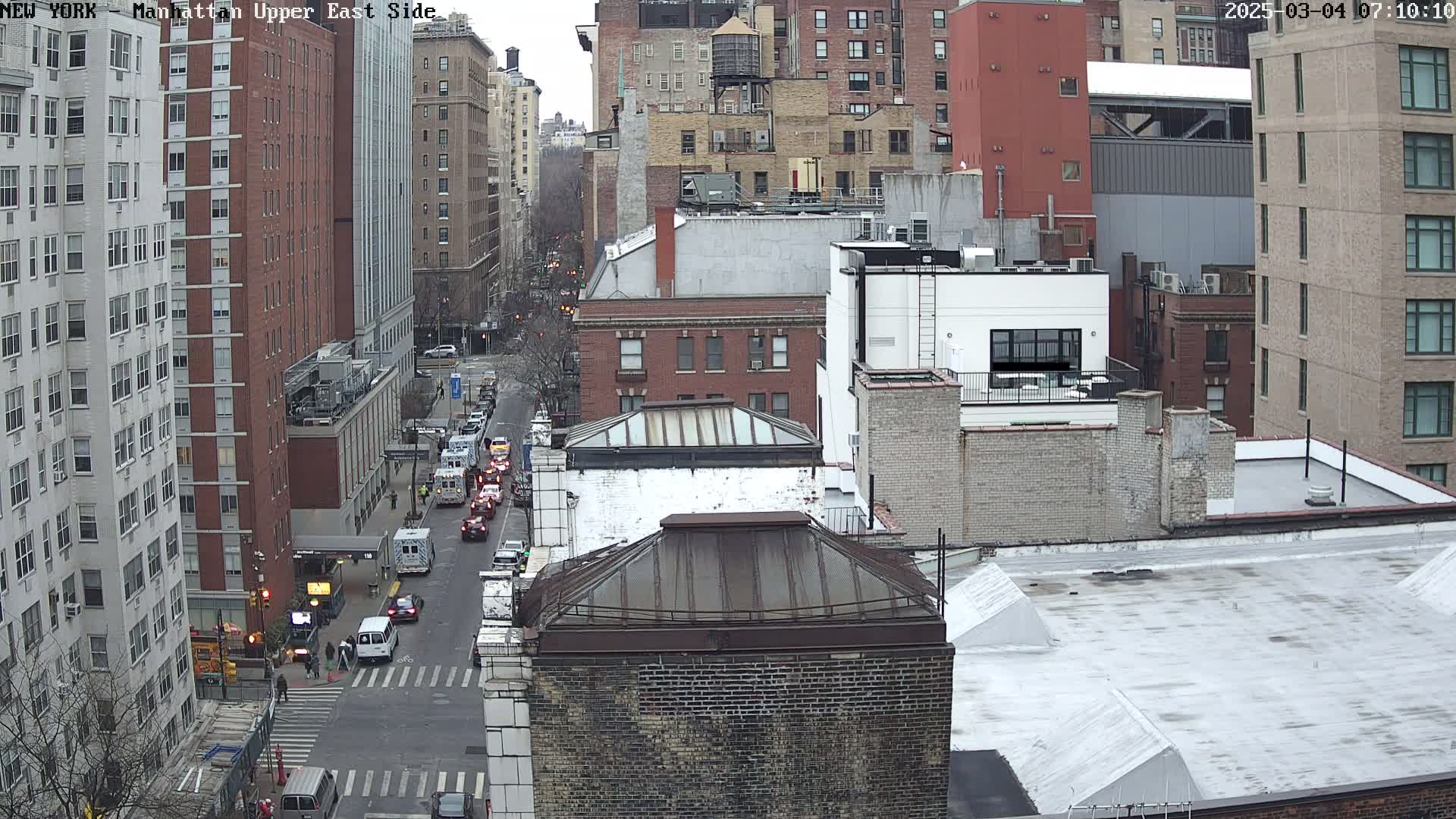 A high-angle, long shot of a city street scene on an overcast day, showing several multi-story buildings and a street with cars.