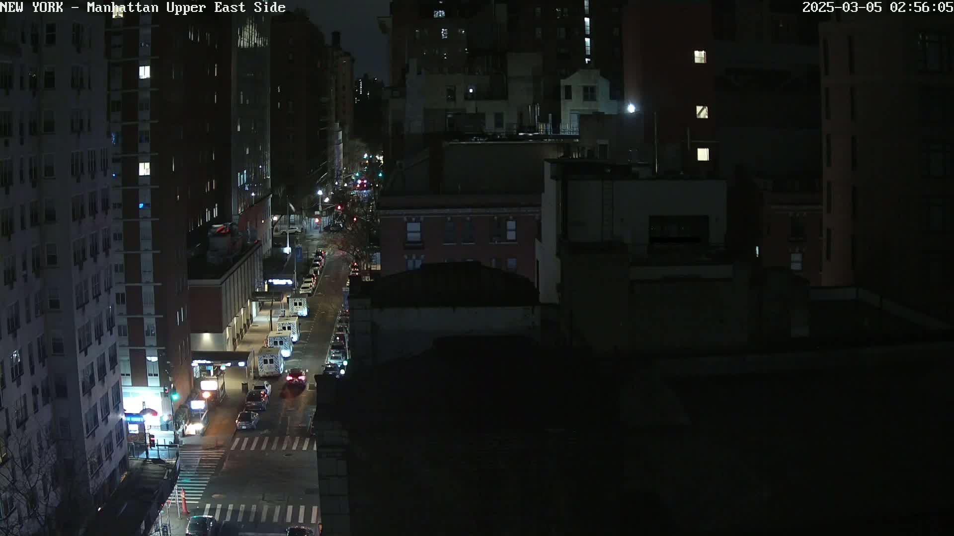 A nighttime, aerial view shows a city street with sparsely parked cars under dark, clear skies.