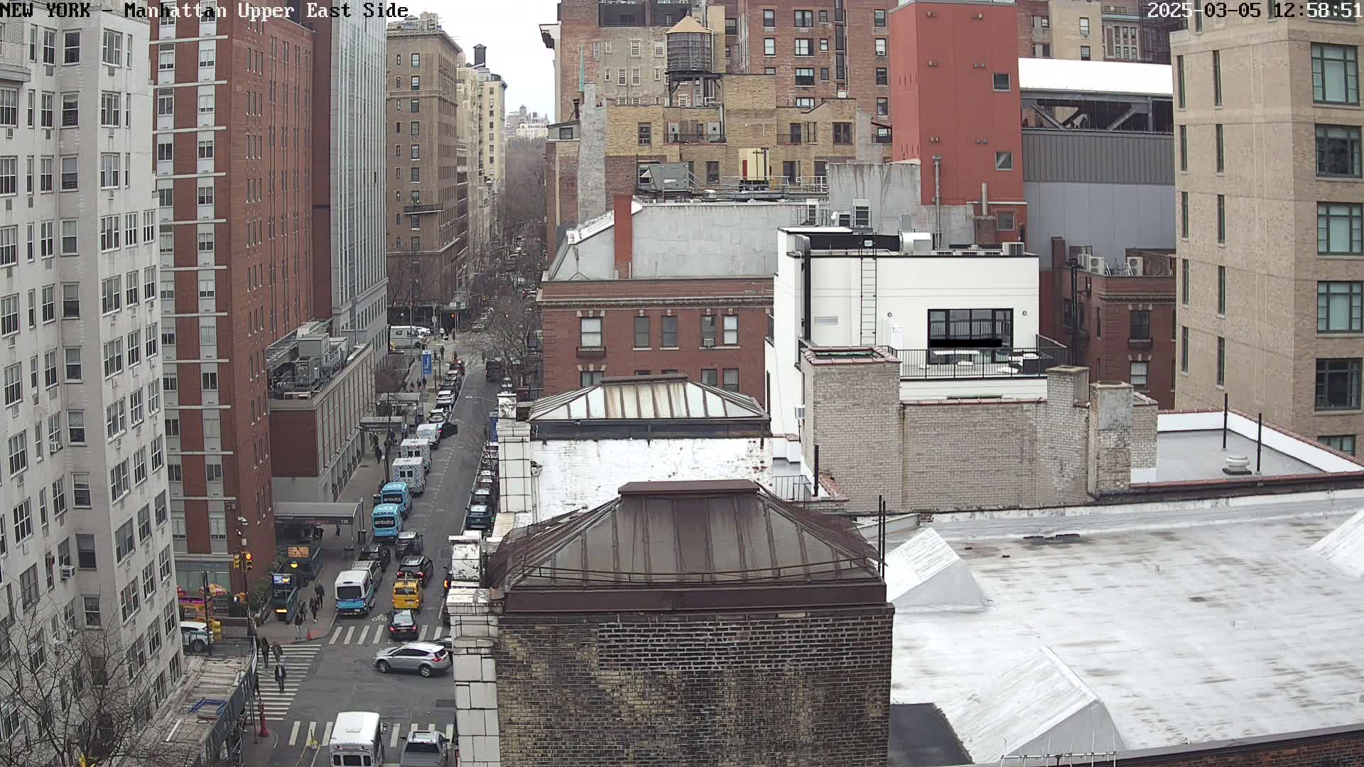 A high-angle, long shot of a city street scene on an overcast day, showing several multi-story buildings and a street with cars and vans.