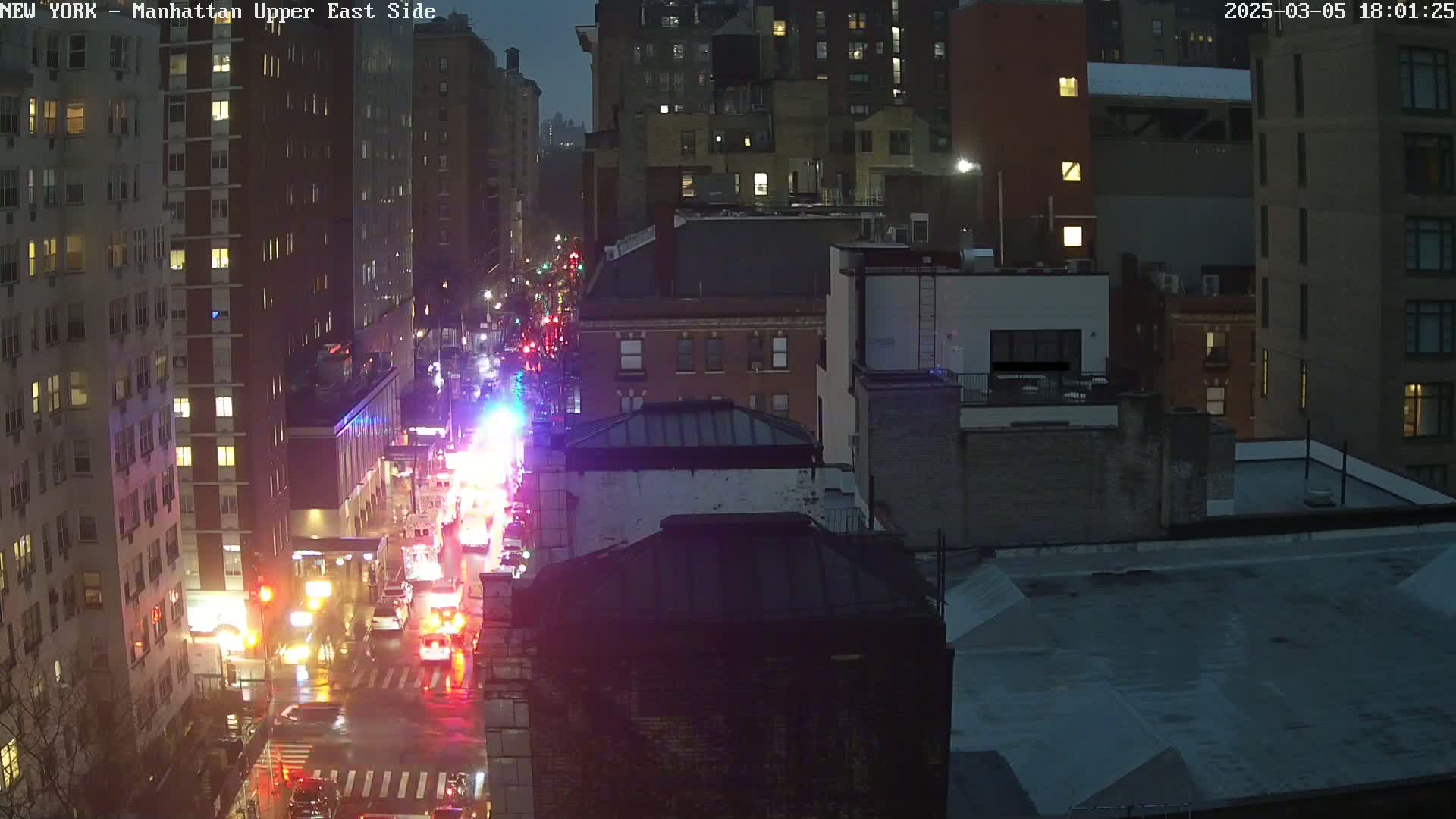 A nighttime view of a rain-slicked city street with emergency vehicles and buildings illuminated by lights.