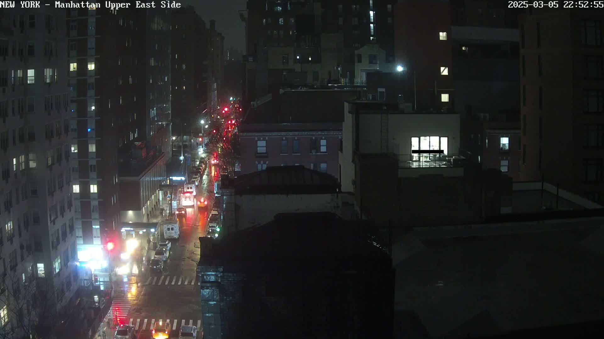 A rain-slicked city street at night, viewed from above, shows several cars driving past dark buildings with illuminated windows.