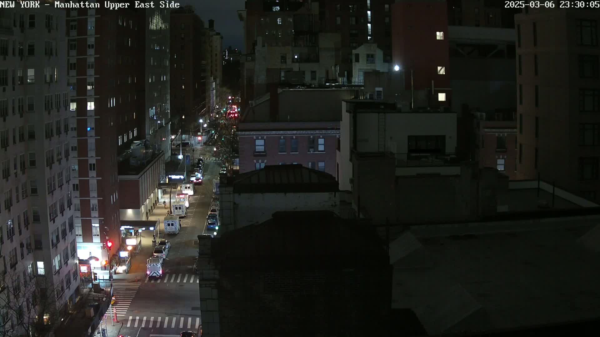 A nighttime, high-angle view of a city street with several vehicles, including emergency vehicles, and numerous buildings under a dark sky.