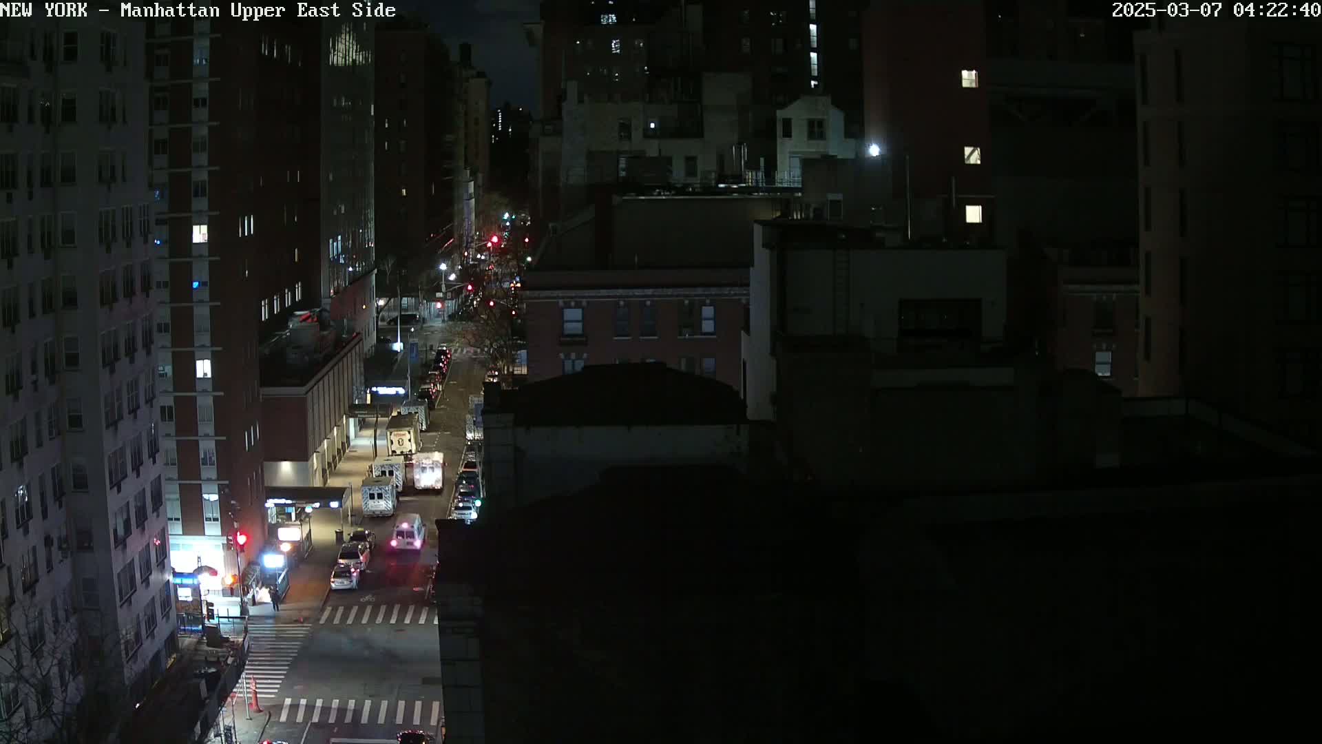 A nighttime, high-angle view of a city street with several vehicles, including emergency vehicles, stopped at an intersection between tall buildings.