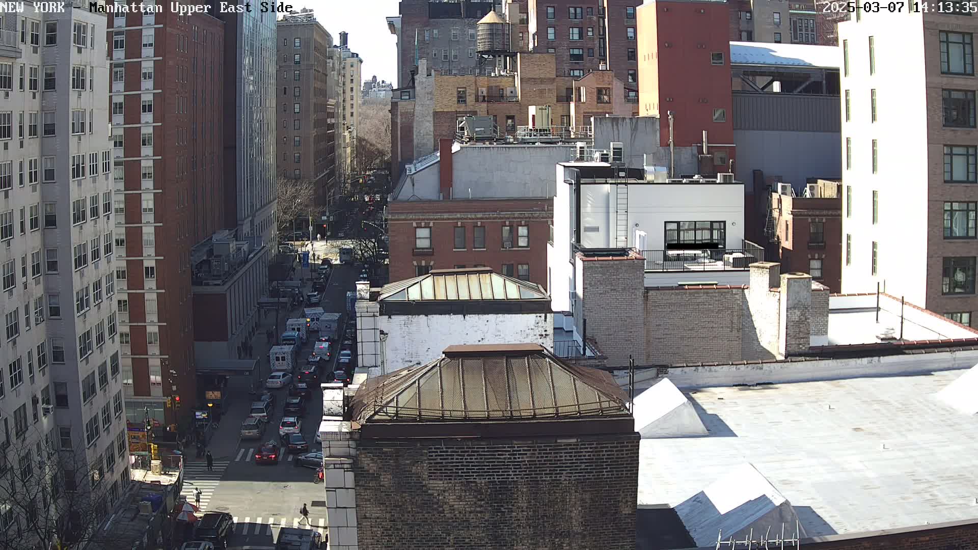 A sunny city street scene shows multiple brick buildings, a few vehicles, and pedestrians on a street below from a high-angle perspective.