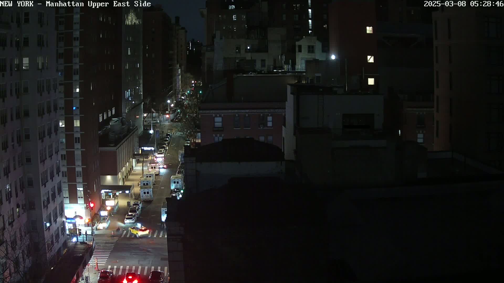 A nighttime aerial view of a city street with several vehicles, including what appear to be emergency vehicles, and tall buildings surrounding it.