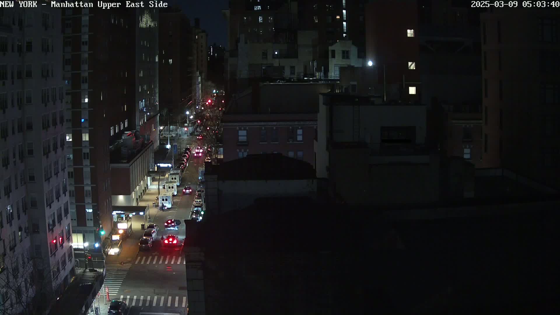 A nighttime, high-angle view of a city street with several cars driving and buildings lining both sides.