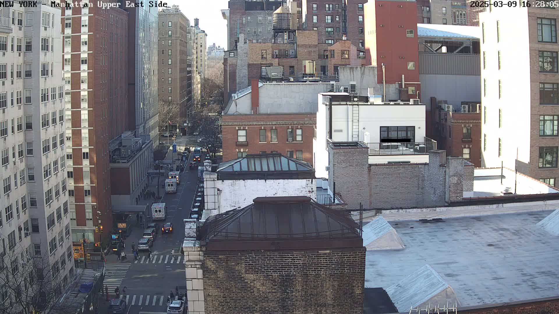 A sunny day view from a rooftop overlooking a city street with several multi-story buildings and some vehicles, including what appears to be an ambulance.