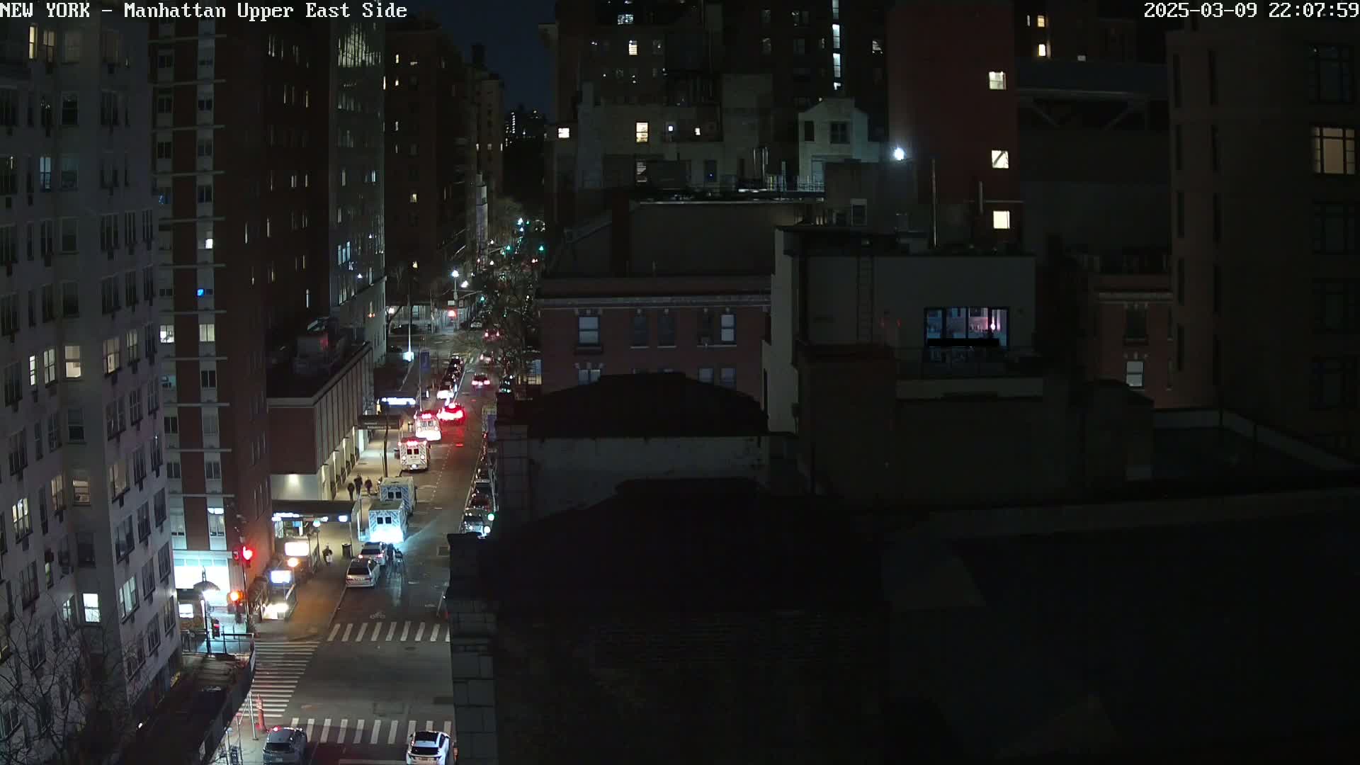 A nighttime, high-angle view of a city street with several vehicles, including emergency vehicles, parked along the side of the road, with buildings lining both sides of the street.