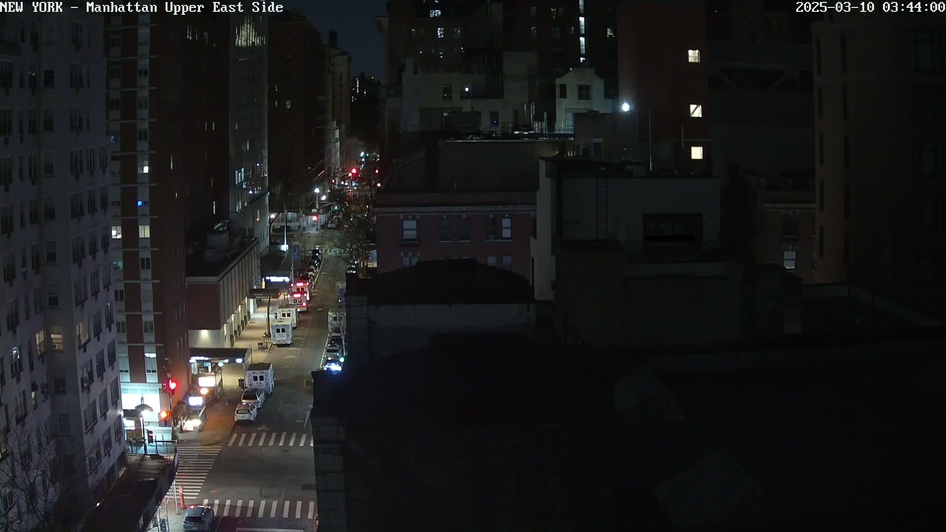 A nighttime, high-angle view of a city street with several emergency vehicles parked along the side of the road, next to tall buildings.