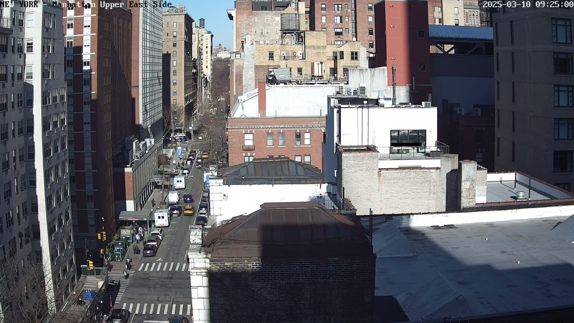 A sunny, high-angle view of a city street, showing several multi-story buildings and cars driving on the road.