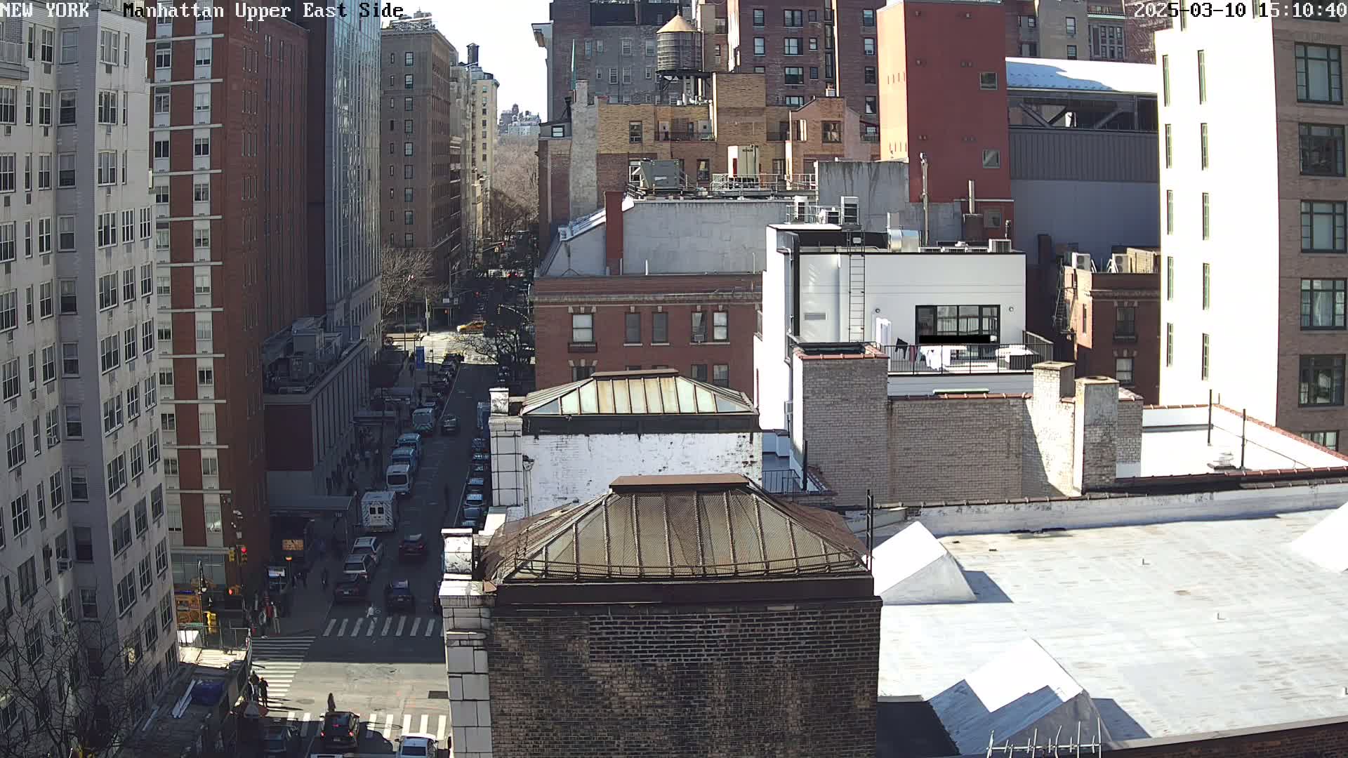 A sunny day view of a city street, with many multi-story buildings, and cars on a cross street.