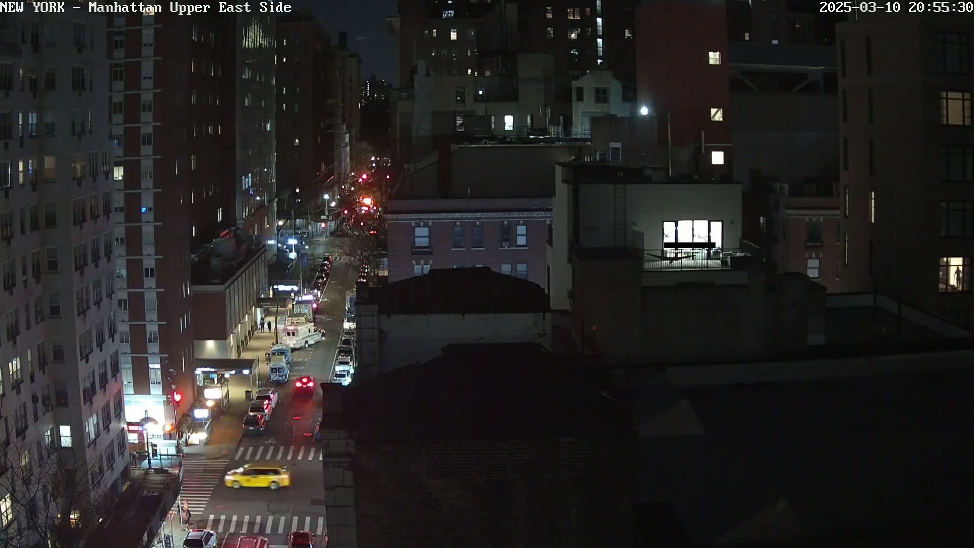 A nighttime, high-angle view of a city street with cars driving on it, bordered by tall buildings.