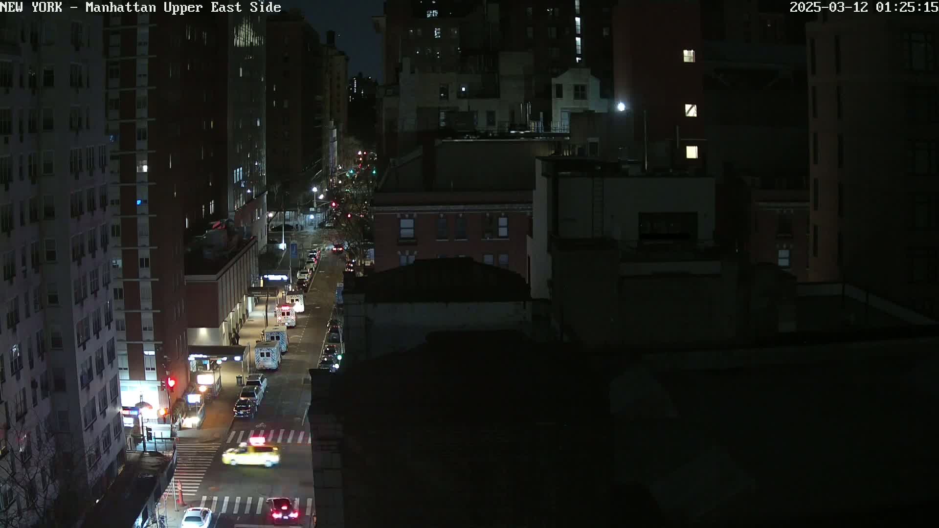 A nighttime, high-angle view of a city street with cars driving past tall buildings under dark, clear skies.
