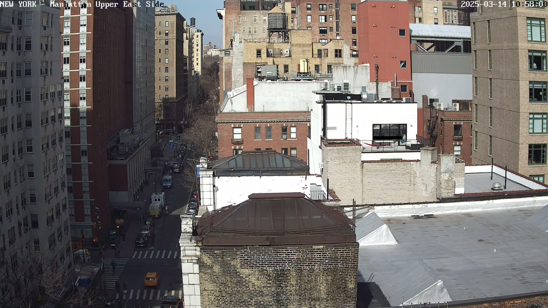 A sunny day view of a city street scene shows numerous brick buildings, a street with cars and pedestrians, and a clear blue sky.