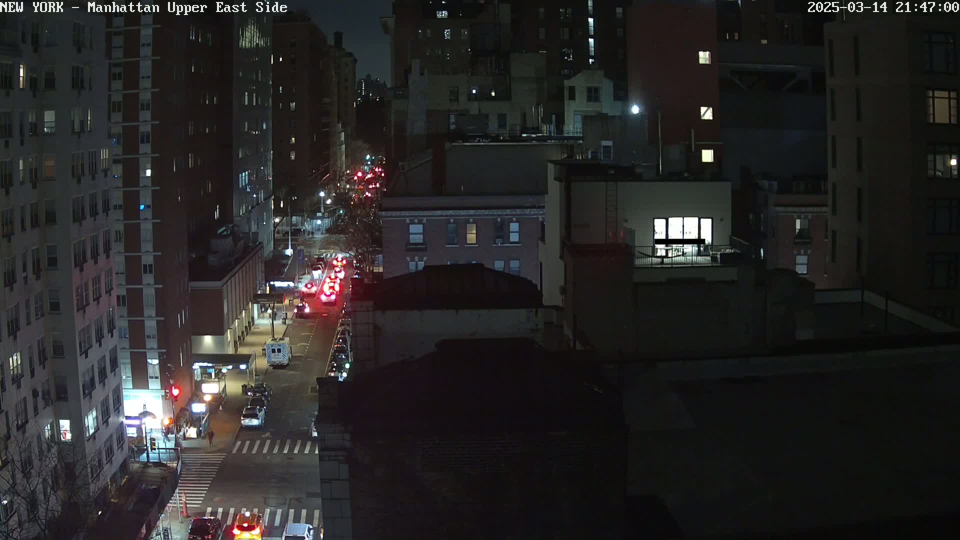 A nighttime, city street scene shows several cars driving on a road between tall buildings under dark, clear skies.