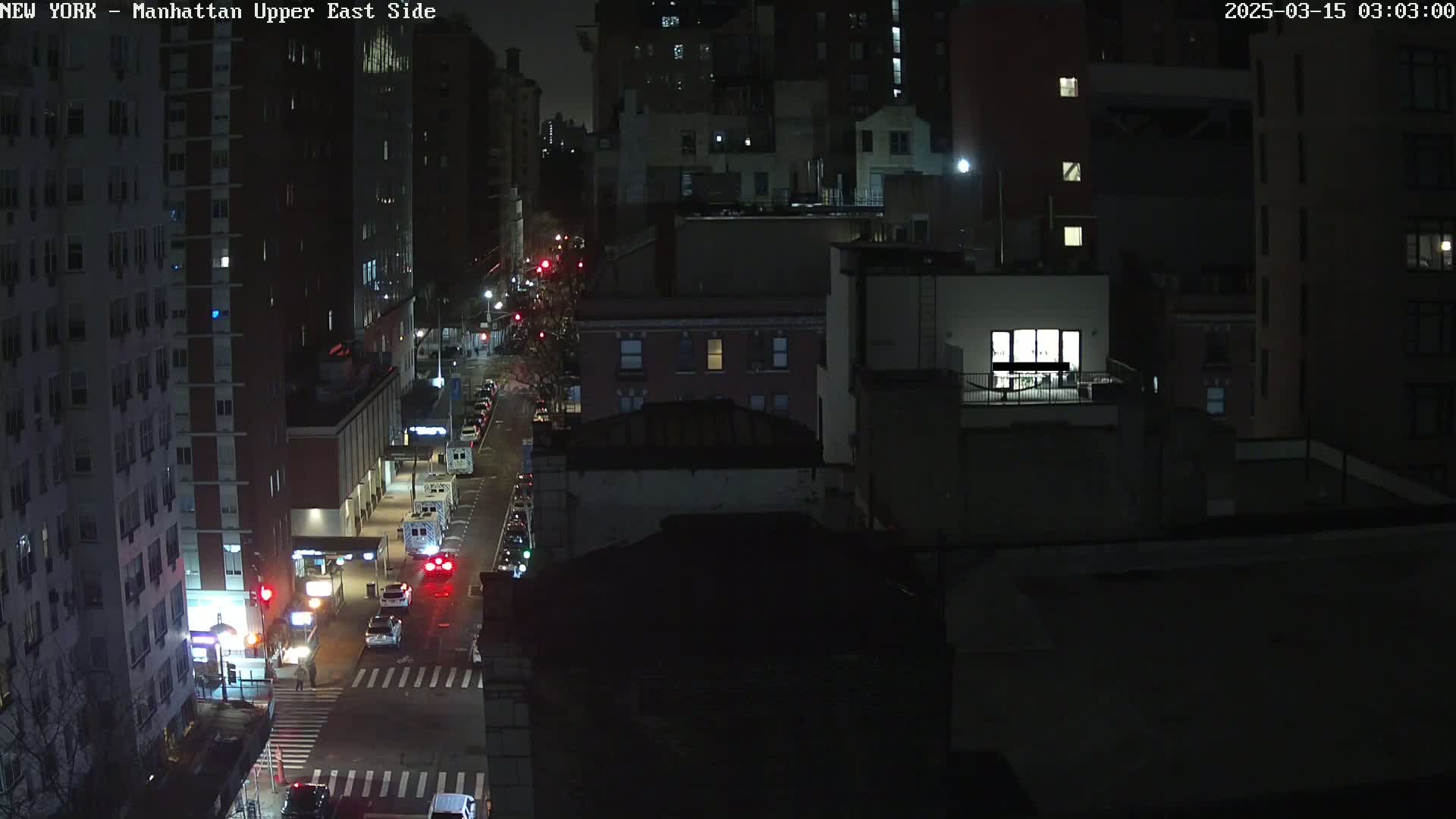 A nighttime, high-angle view of a city street with several cars, buildings, and a few illuminated windows.