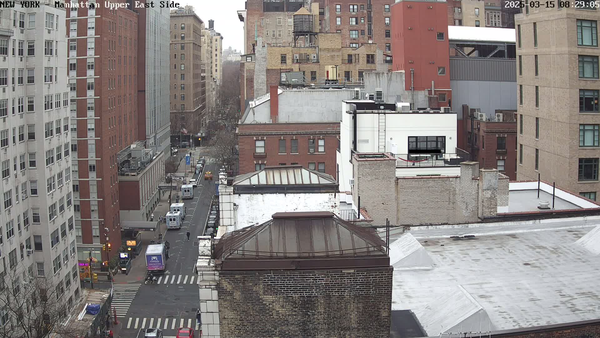 A high-angle view of a city street with several multi-story buildings on an overcast day, showing vehicles on the street and some sparse pedestrian activity.