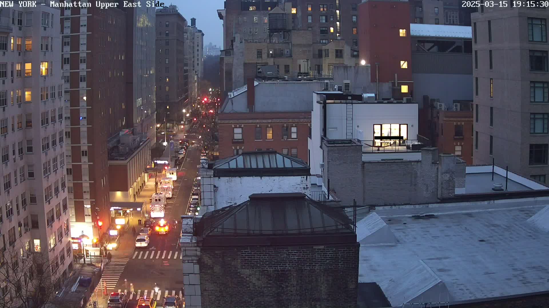 A twilight view of a city street with several multi-story buildings and traffic, including emergency vehicles.