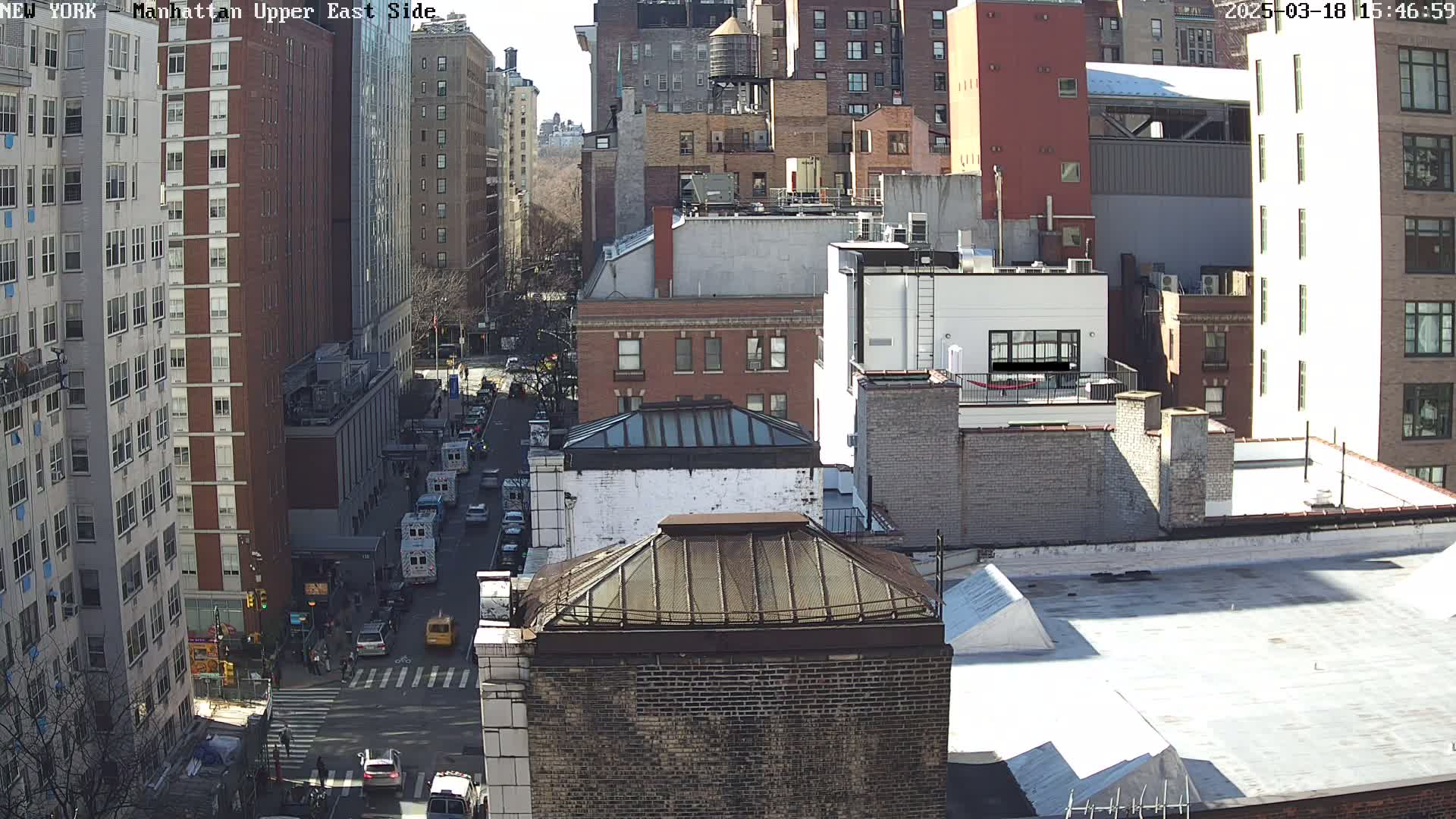 A sunny day view of a city street with several multi-story buildings, cars, and pedestrians.
