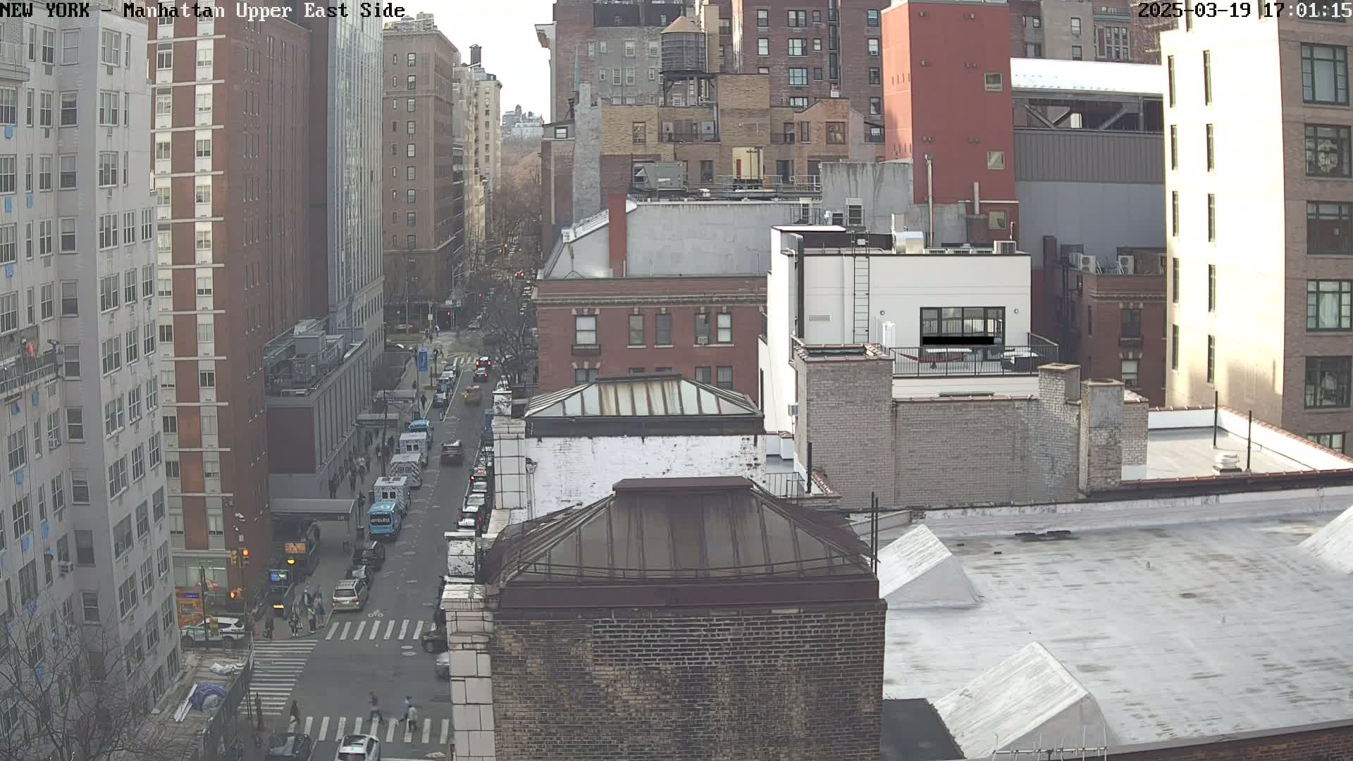 A high-angle, long shot of a city street with several multi-story buildings on a partly sunny day.