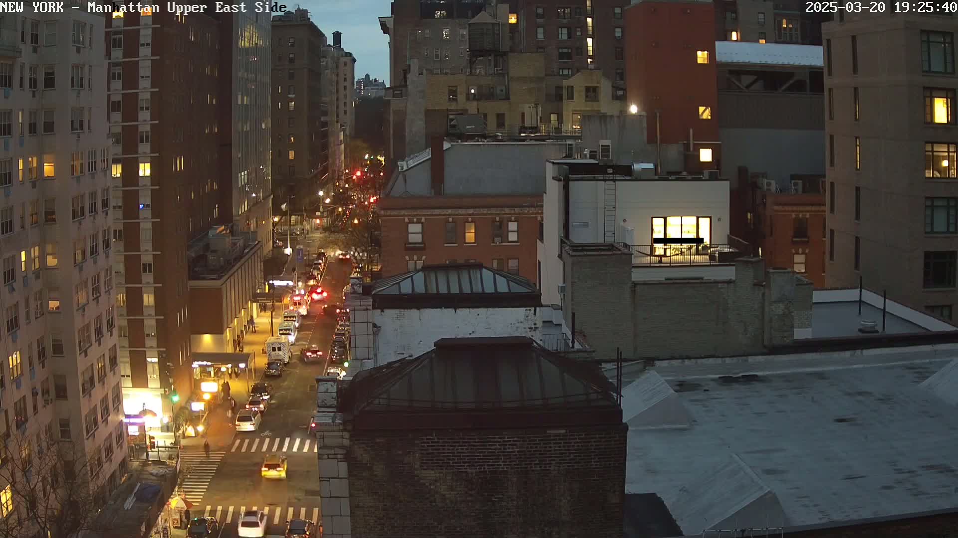 A nighttime aerial view of a city street shows traffic moving along a road lined with tall buildings under a dusky sky.