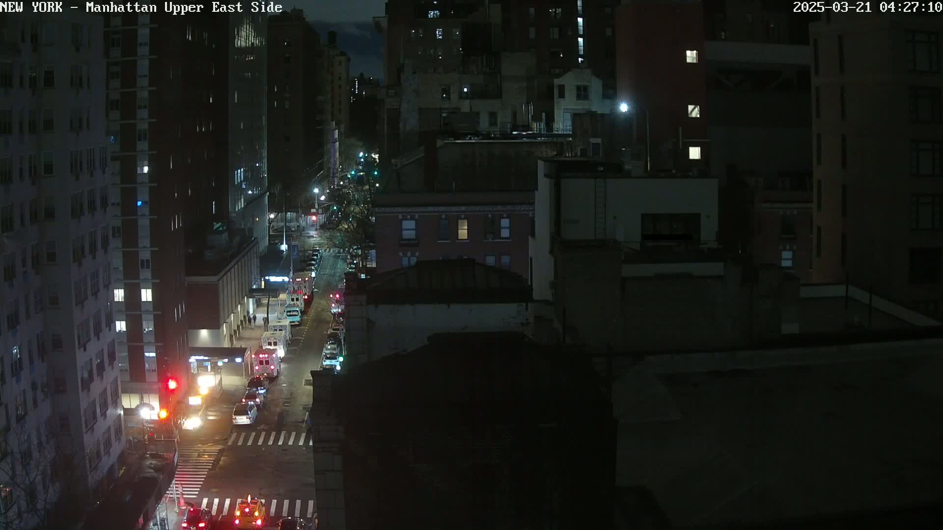 A nighttime, high-angle view of a city street with several emergency vehicles stopped at an intersection, surrounded by dark buildings.