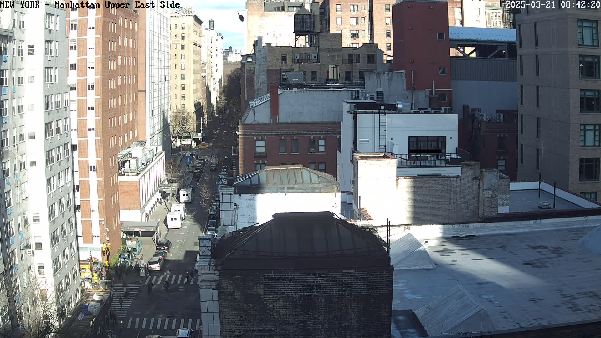 A sunny, high-angle view of a city street scene shows several multi-story buildings, vehicles, and pedestrians, with shadows cast on the rooftops and street below.