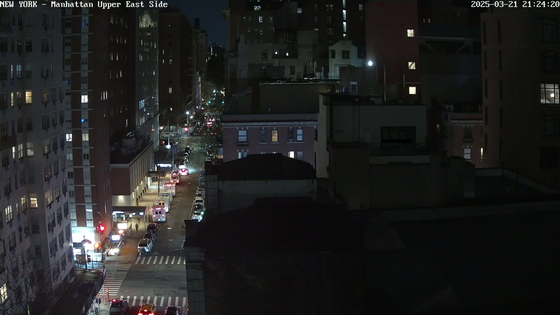 A nighttime, high-angle view of a city street with several buildings and cars, under dark conditions.