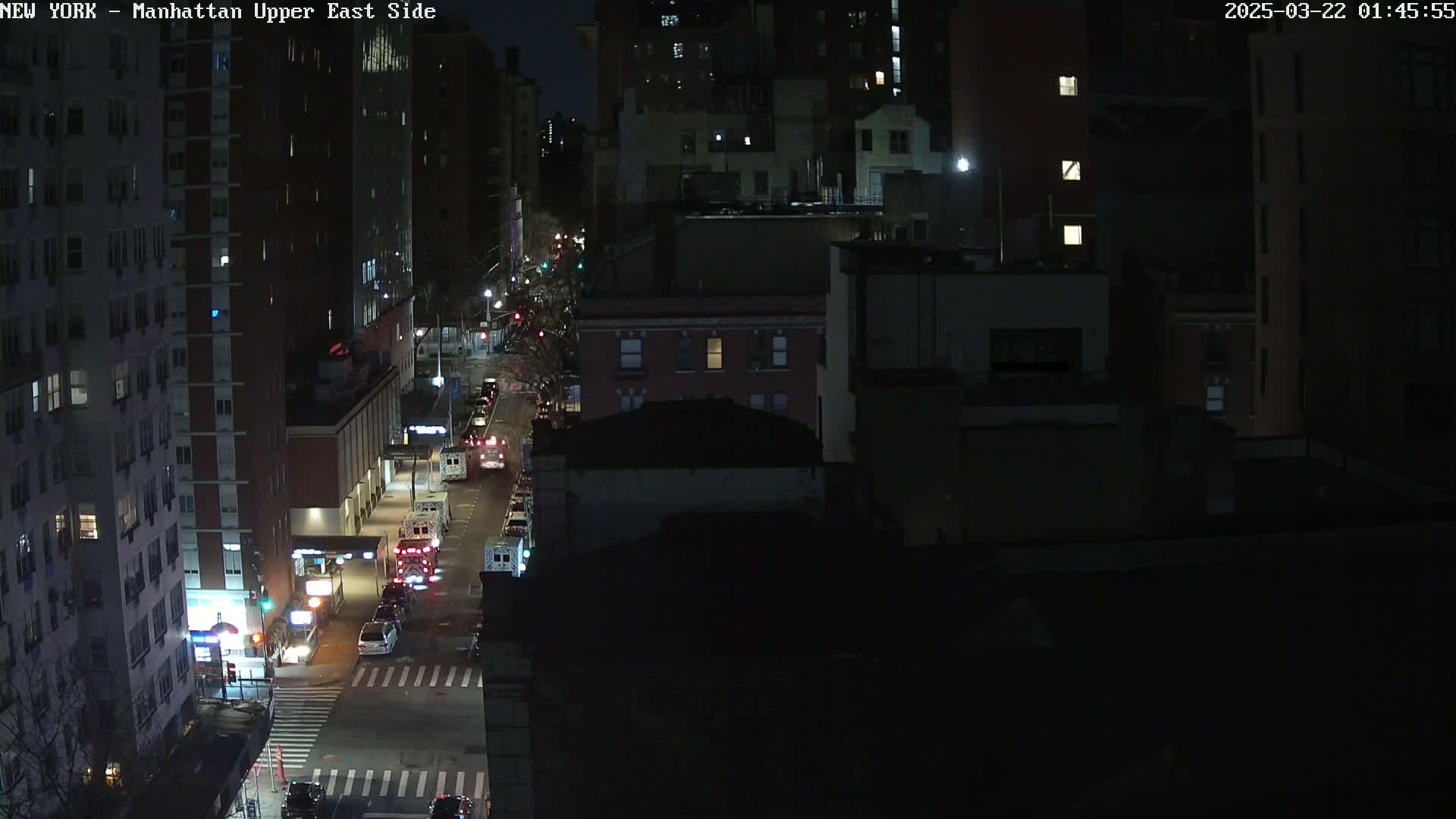 A nighttime, high-angle view of a city street with several emergency vehicles parked along the side of the road in between tall buildings.