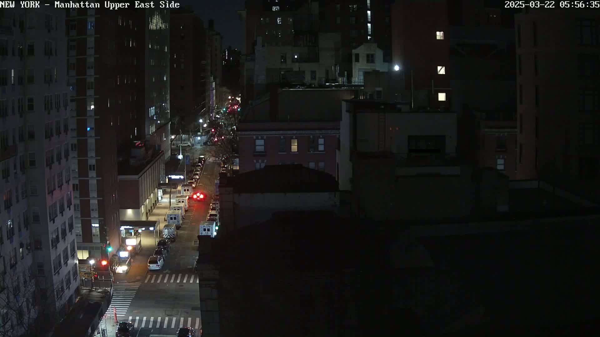 A nighttime aerial view of a city street shows several vehicles, including emergency vehicles, parked along the side of the road, with tall buildings surrounding the street.