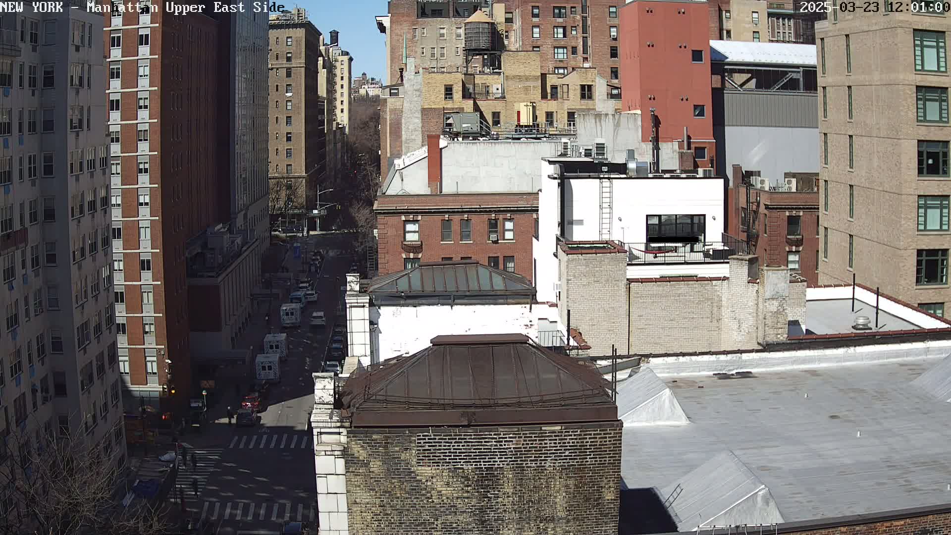 A sunny day view of a city street, showing multiple brick buildings, rooftops, and vehicles on the street below.