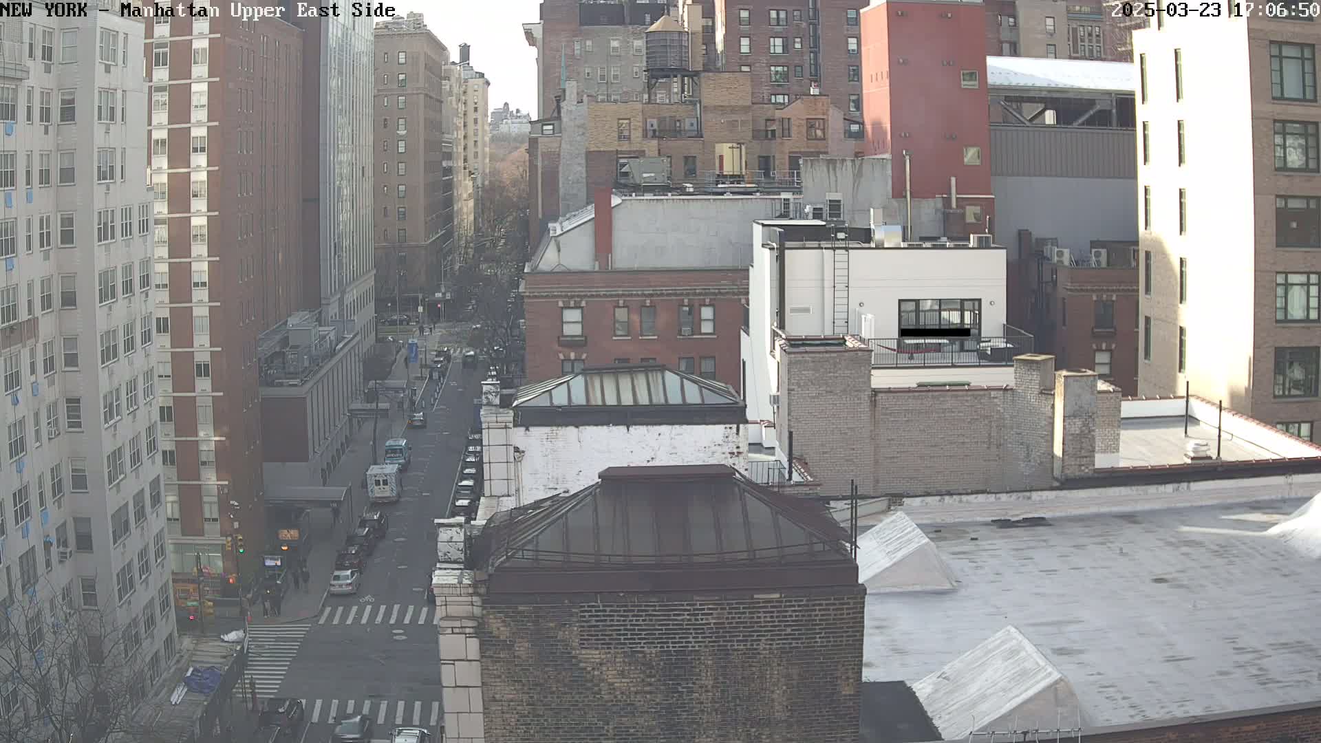 A high-angle view of a city street on a mostly clear day shows several multi-story brick buildings and a few cars parked along the street.