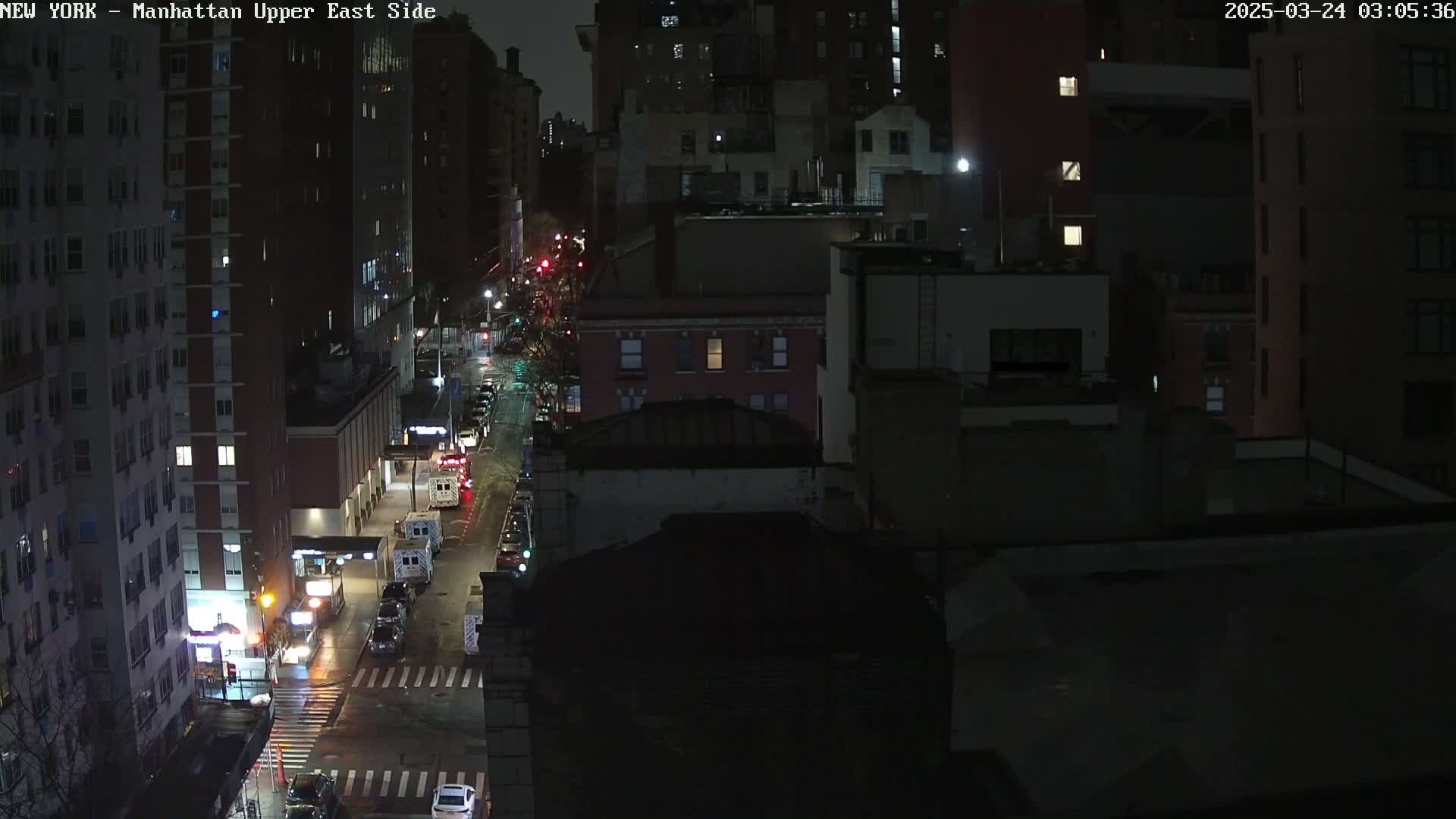 A nighttime, high-angle view of a city street with several vehicles, including emergency vehicles, and tall buildings under dark, seemingly clear skies.