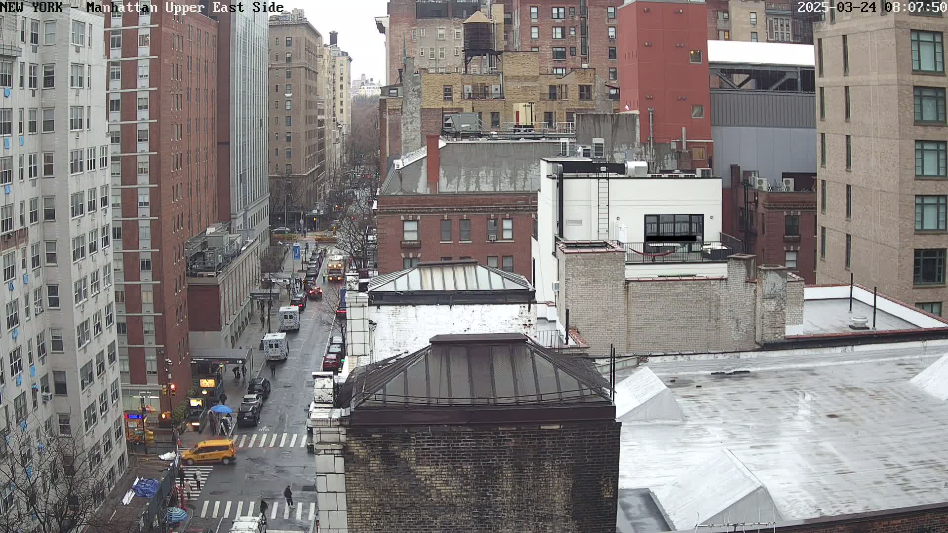 A rain-slicked city street scene shows several multi-story brick buildings, vehicles, and pedestrians at a street intersection.