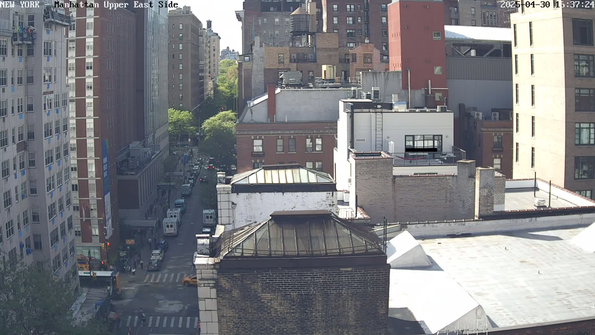 A sunny cityscape view of several brick buildings, a street with vehicles, and a few pedestrians.