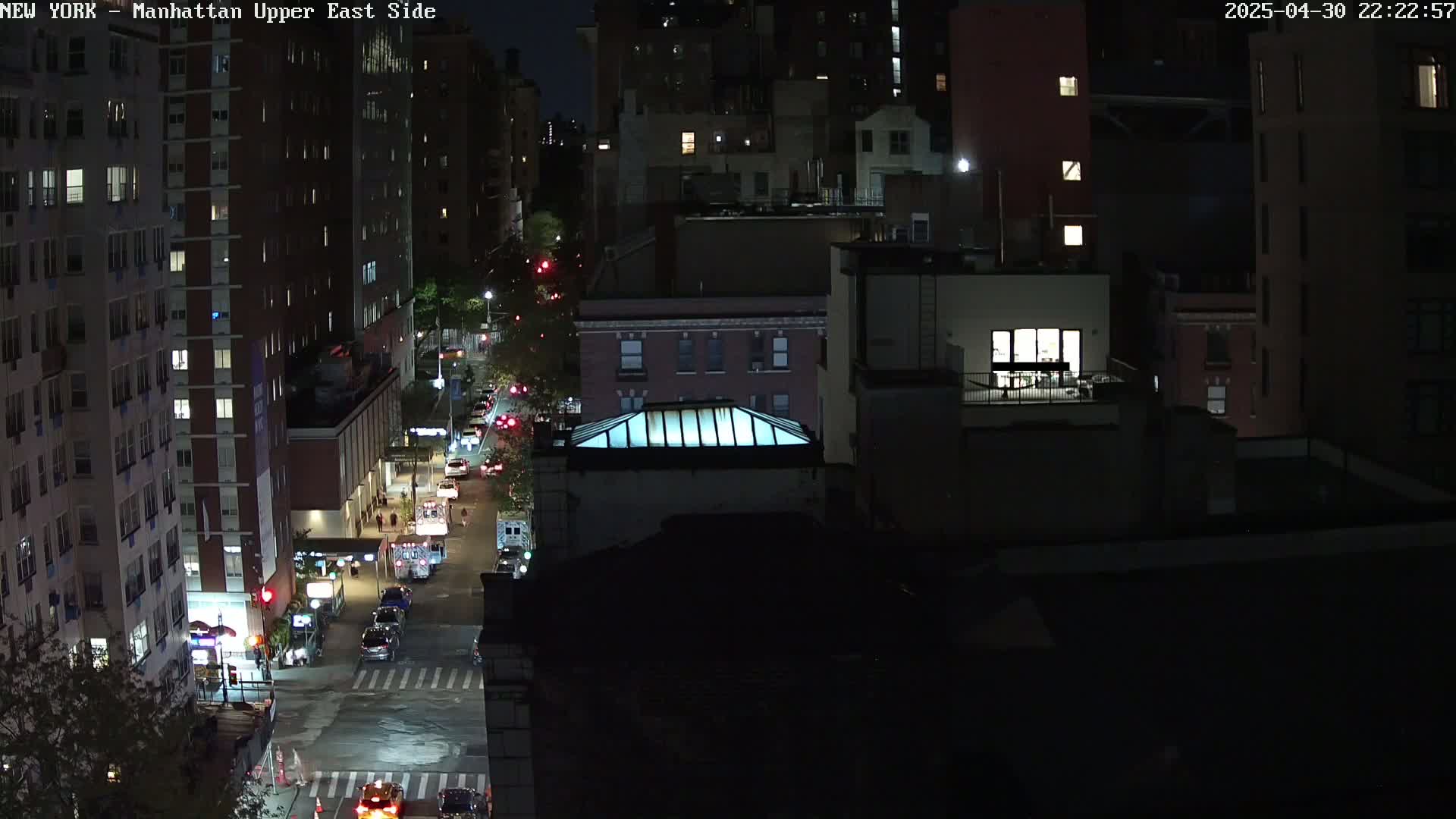 A nighttime aerial view of a city street with several multi-story buildings, showing cars and some dimly lit areas.