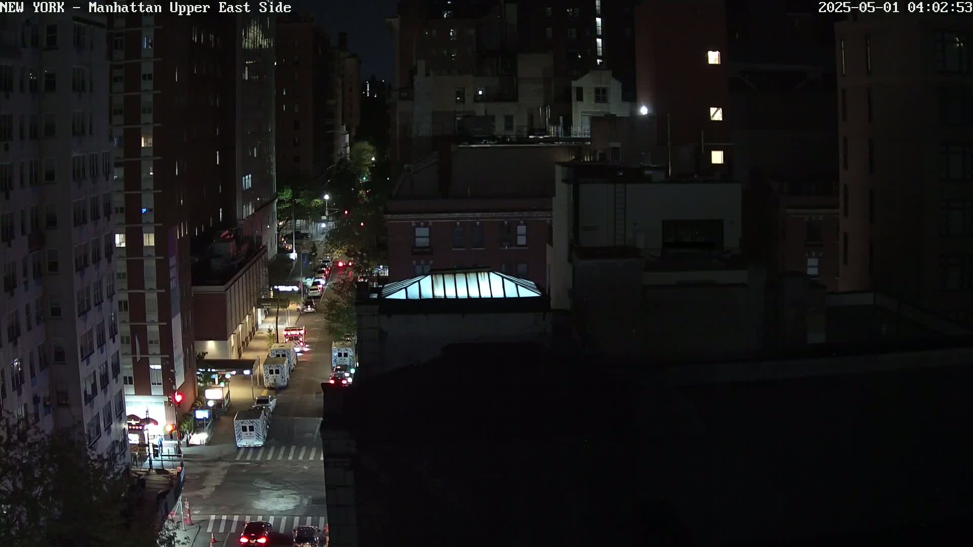 A nighttime aerial view shows several city blocks with dimly lit buildings and a street with several emergency vehicles.