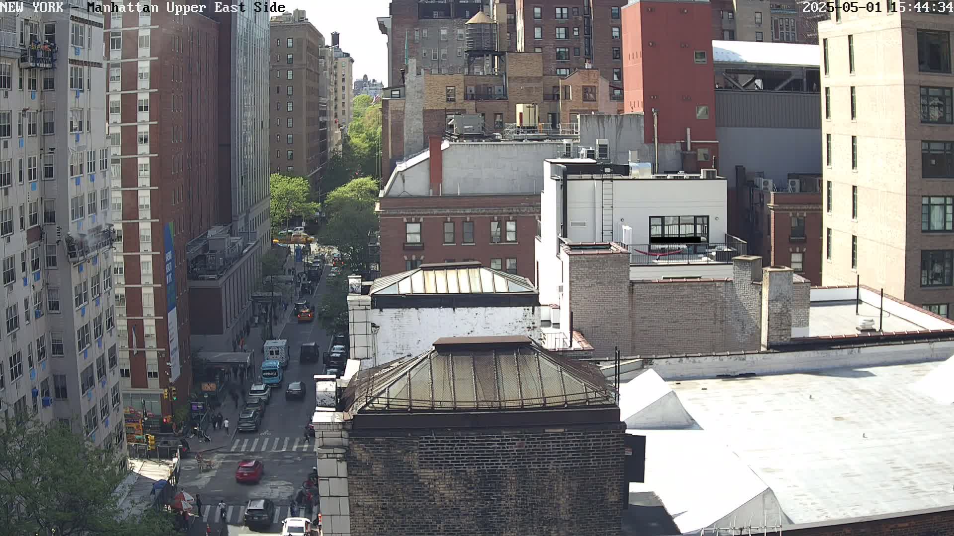A sunny day view of a city street with several multi-story buildings, cars, and pedestrians.
