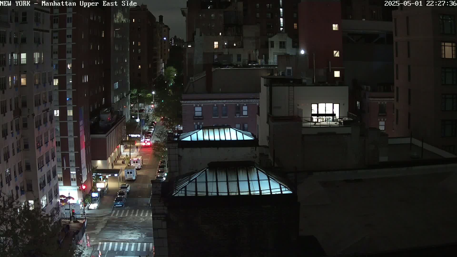 A nighttime aerial view of a city street shows several multi-story buildings, vehicles, and two glass-topped structures, under dark conditions.