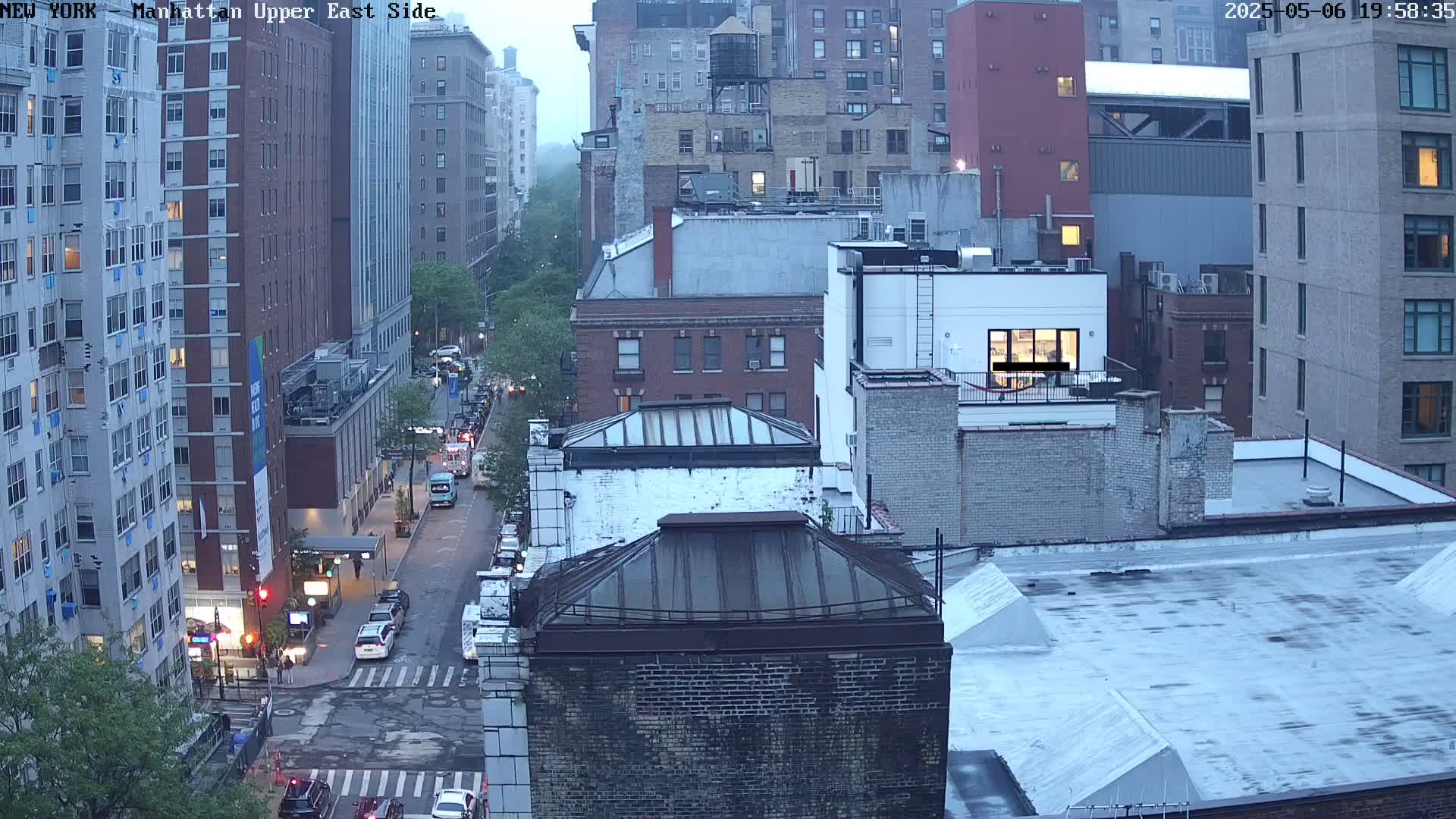 A high-angle view of a city street at dusk, showing several multi-story buildings and cars, under an overcast sky.