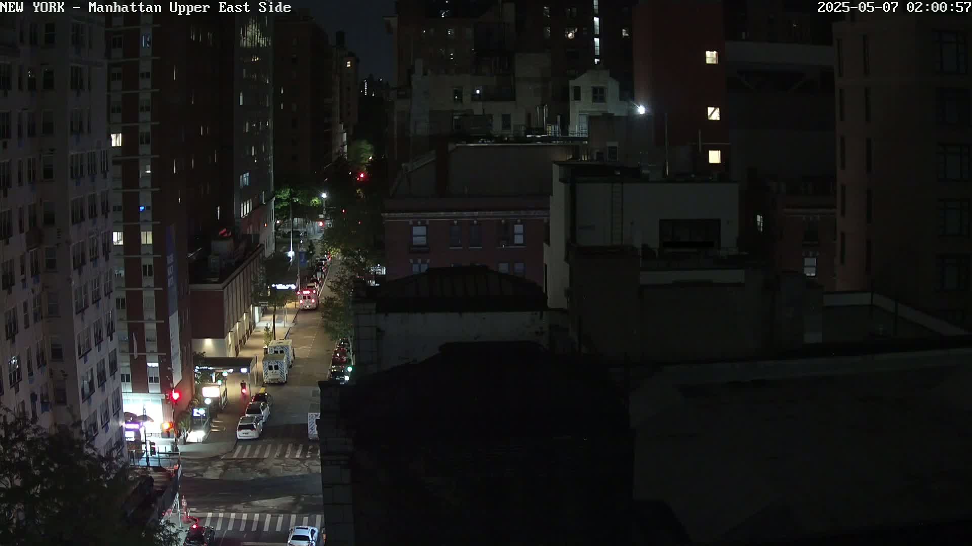 A nighttime, high-angle view of a city street shows several vehicles parked and driving on a road between tall buildings.