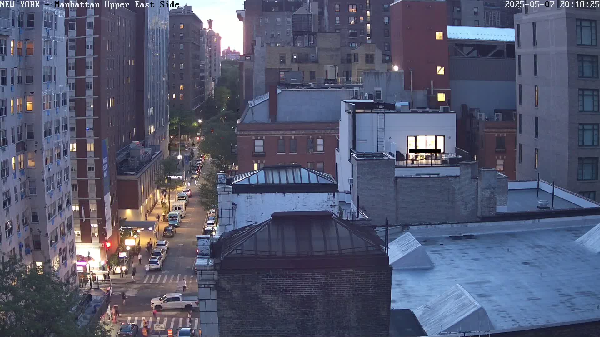 A twilight view of a city street shows several multi-story buildings, cars parked along the side of the road, and pedestrians walking on the sidewalk.