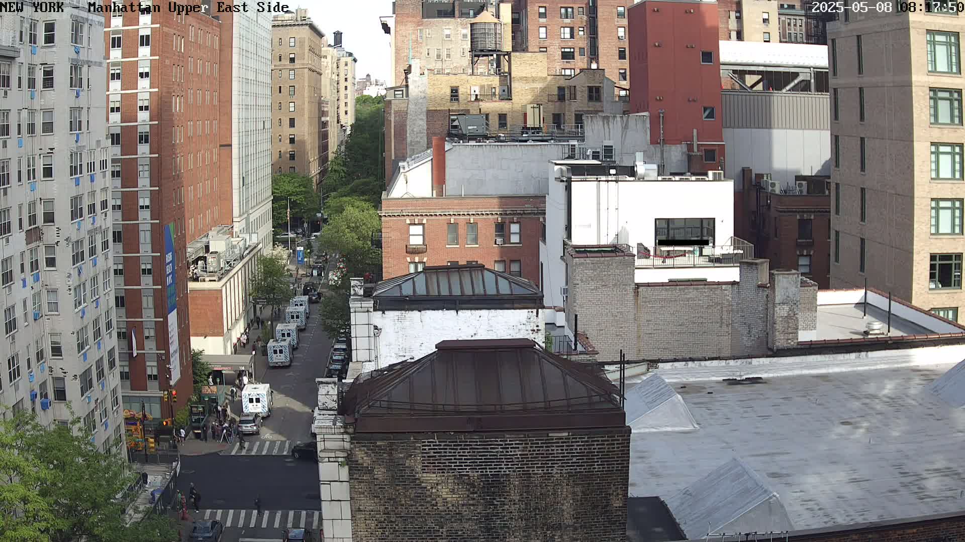A high-angle view of a city street scene on a sunny day, showing numerous brick buildings, parked vehicles, and a few pedestrians.