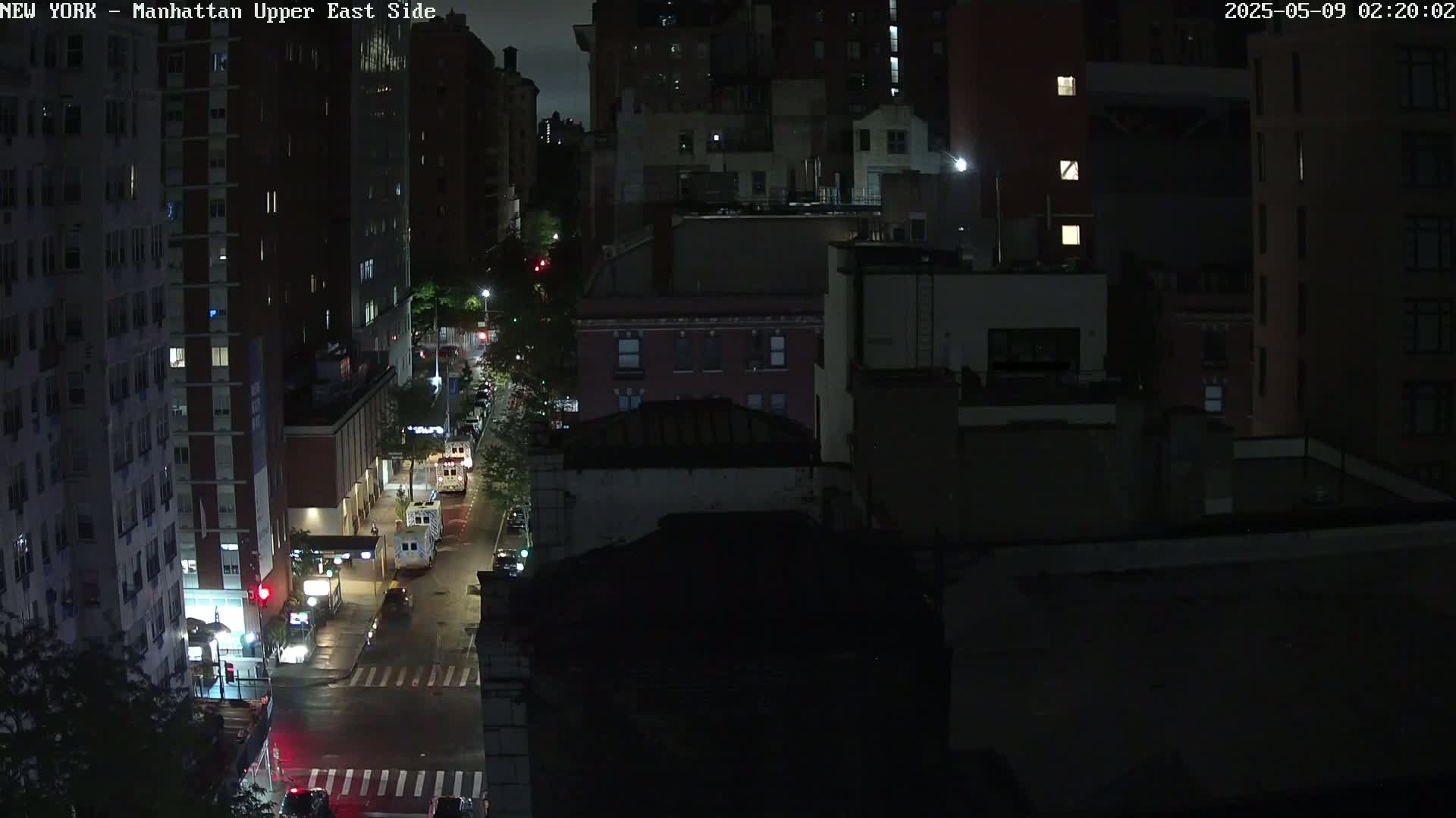 A nighttime, high-angle view of a city street with several vehicles, illuminated by streetlights, bordered by dark buildings.