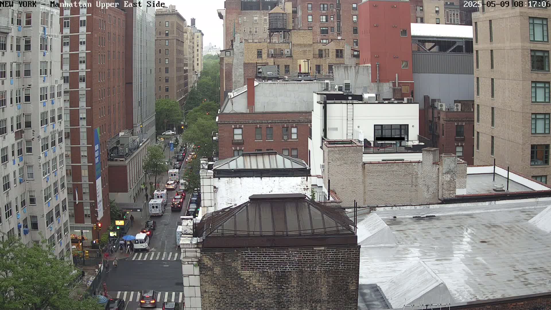 An overcast day reveals a city street scene with several multi-story buildings, some vehicles on the street, and a few pedestrians at a crosswalk.